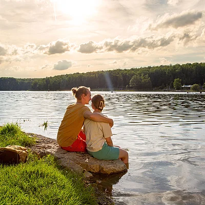 Zwei Personen sitzen am Ufer eines Sees auf einem Stein, eine hat den Arm um die andere gelegt.
