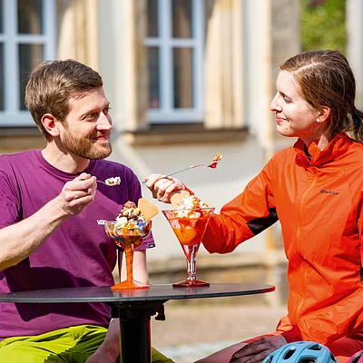 Zwei Personen sitzen draußen an einem Tisch und essen Eis aus Gläsern bei Sonnenschein.
