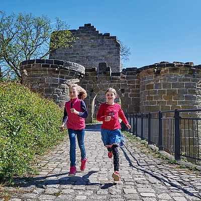 Zwei Mädchen laufen auf einem gepflasterten Weg vor einer alten Steinburg bei klarem Himmel.