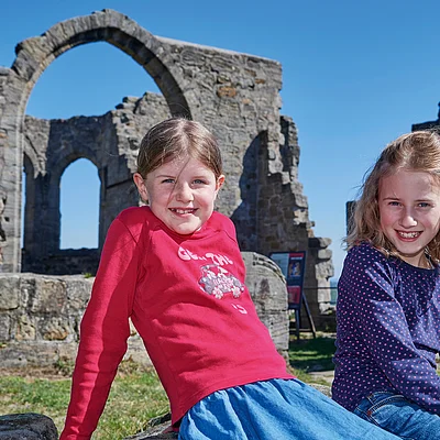 Zwei Mädchen sitzen vor einer alten Steinruine mit Bögen bei klarem Himmel.