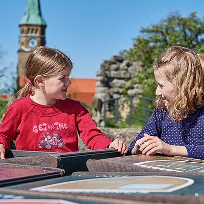 Zwei Mädchen sitzen draußen an einem Tisch mit Informationstafeln und schauen sich an.