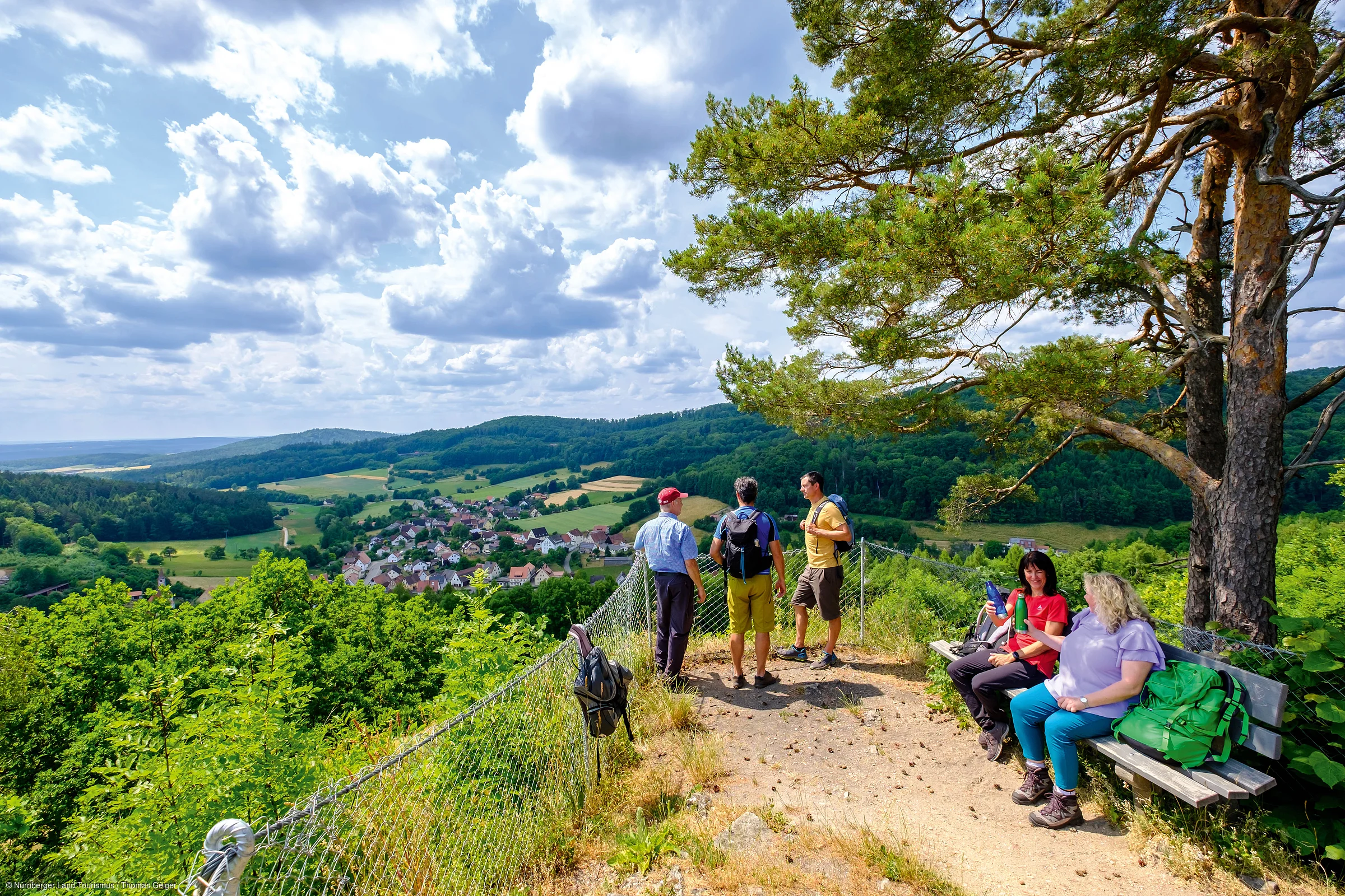 Wandergruppe auf Aussichtsplattform mit Blick auf Tal, Dorf und bewaldete Hügel unter bewölktem Himmel.