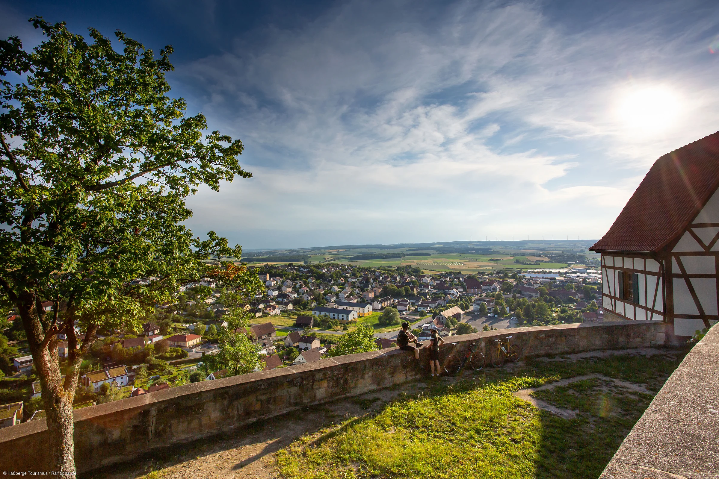 Blick von einer Anhöhe auf eine Stadt mit Häusern, zwei Personen an einer Mauer und einem Fachwerkhaus rechts.