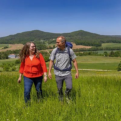 Zwei Personen wandern auf einer grünen Wiese mit Hügeln und Feldern im Hintergrund bei klarem Himmel.