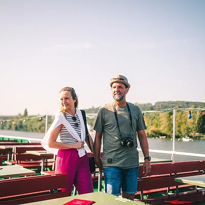 Paar geht auf Terrasse mit roten Bänken und grünen Tischen am Fluss bei Sonnenschein spazieren.