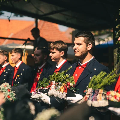 Junge Männer in traditionellen Trachten mit roten Westen halten geschmückte Hüte bei einer Veranstaltung im Freien.
