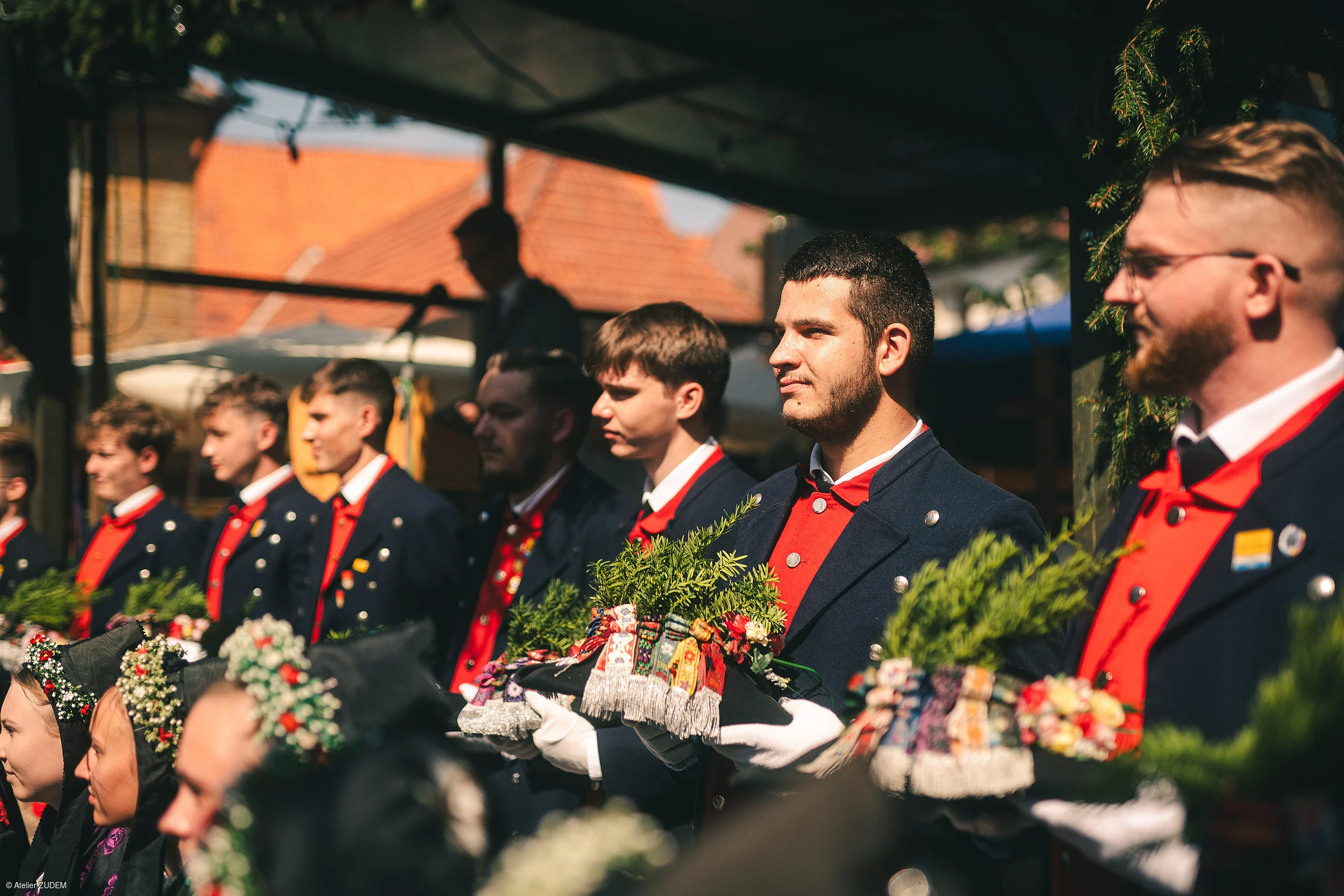 Junge Männer in traditionellen Trachten mit roten Westen halten geschmückte Hüte bei einer Veranstaltung im Freien.