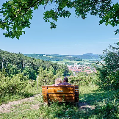 Paar sitzt auf einer Holzbank auf einem Hügel mit Blick auf Wald, Tal und Dorf im Hintergrund.