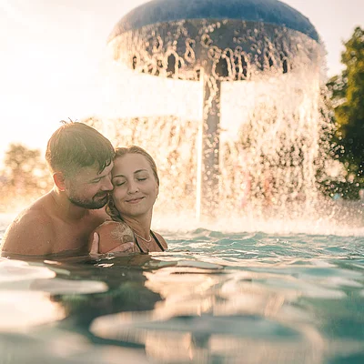 Paar umarmt sich im Wasser vor einem großen Pilzbrunnen bei Sonnenuntergang im Freien.