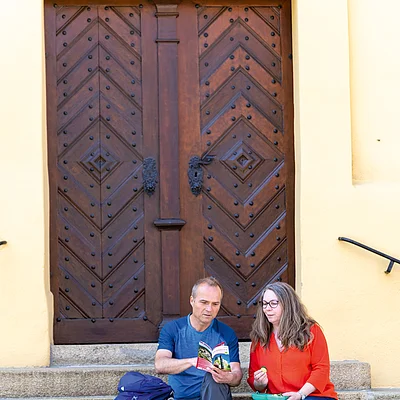 Zwei Personen mit Rucksäcken sitzen auf Steinstufen vor einer großen Holztür und essen Snack.
