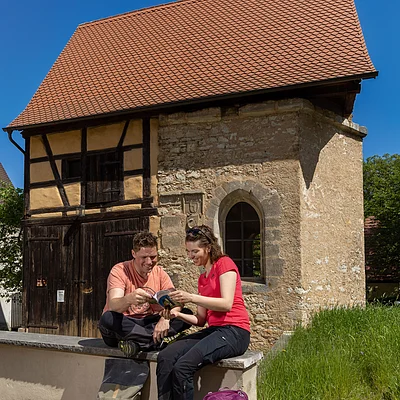 Zwei Personen sitzen vor einem Fachwerkhaus und schauen gemeinsam in einen Reiseführer bei sonnigem Wetter.