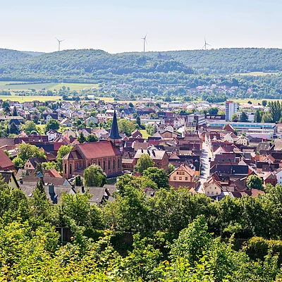 Blick auf eine Kleinstadt mit Kirche, umgeben von Bäumen, Feldern und bewaldeten Hügeln mit Windrädern im Hintergrund.