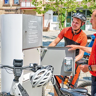 Zwei Männer an einer geöffneten Fahrrad-Ladestation im Freien, ein Mann trägt Fahrradhelm und orange-schwarzes Trikot.
