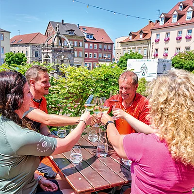 Vier Personen stoßen mit Weingläsern an, sitzen an einem Holztisch im Freien in einer Stadt mit historischen Gebäuden.
