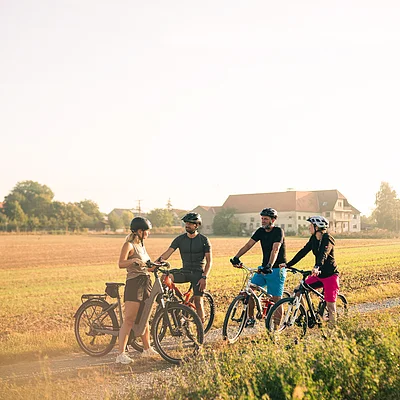 Vier Personen mit Fahrradhelmen stehen mit ihren Fahrrädern auf einem Feldweg in ländlicher Umgebung.