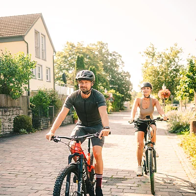 Zwei Personen mit Helmen fahren bei Sonnenschein auf gepflastertem Weg mit Fahrrädern an Häusern und Bäumen vorbei.