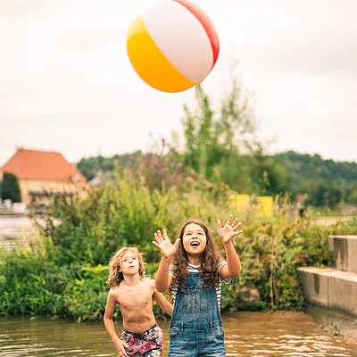 Zwei Kinder spielen mit einem bunten Ball im flachen Wasser eines Teichs oder Sees.