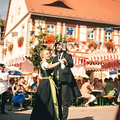 Paar in traditioneller Tracht tanzt auf einem Fest vor einem Fachwerkhaus mit bunten Fahnen und Marktständen.