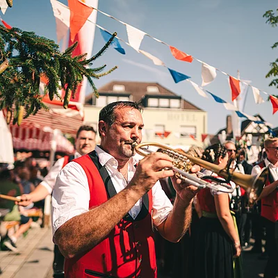 Mann in roter Weste spielt Trompete bei Kirchweih mit Menschen und Girlanden im Hintergrund.