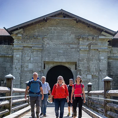 Fünf Personen mit Rucksäcken gehen auf einer Holzbrücke vor einem alten steinernen Torhaus bei Sonnenschein.