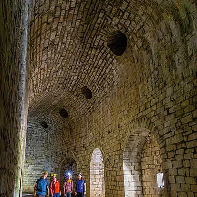 Vier Personen mit Helmen stehen in einem großen, gewölbten Raum aus hellen Steinen mit mehreren Rundbögen.