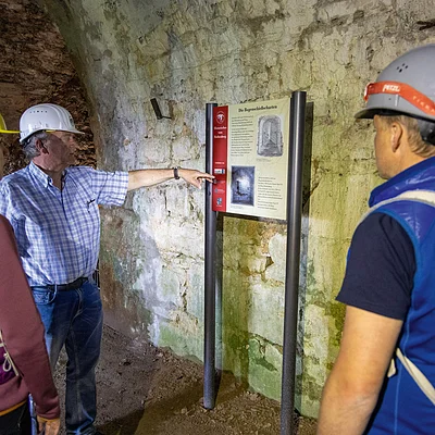 Drei Personen mit Schutzhelmen betrachten eine Informationstafel in einem Tunnel mit steinernen Wänden.