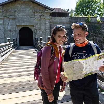Zwei Wanderer mit Rucksäcken lesen eine Karte auf einer Holzbrücke vor einem steinernen Tor.