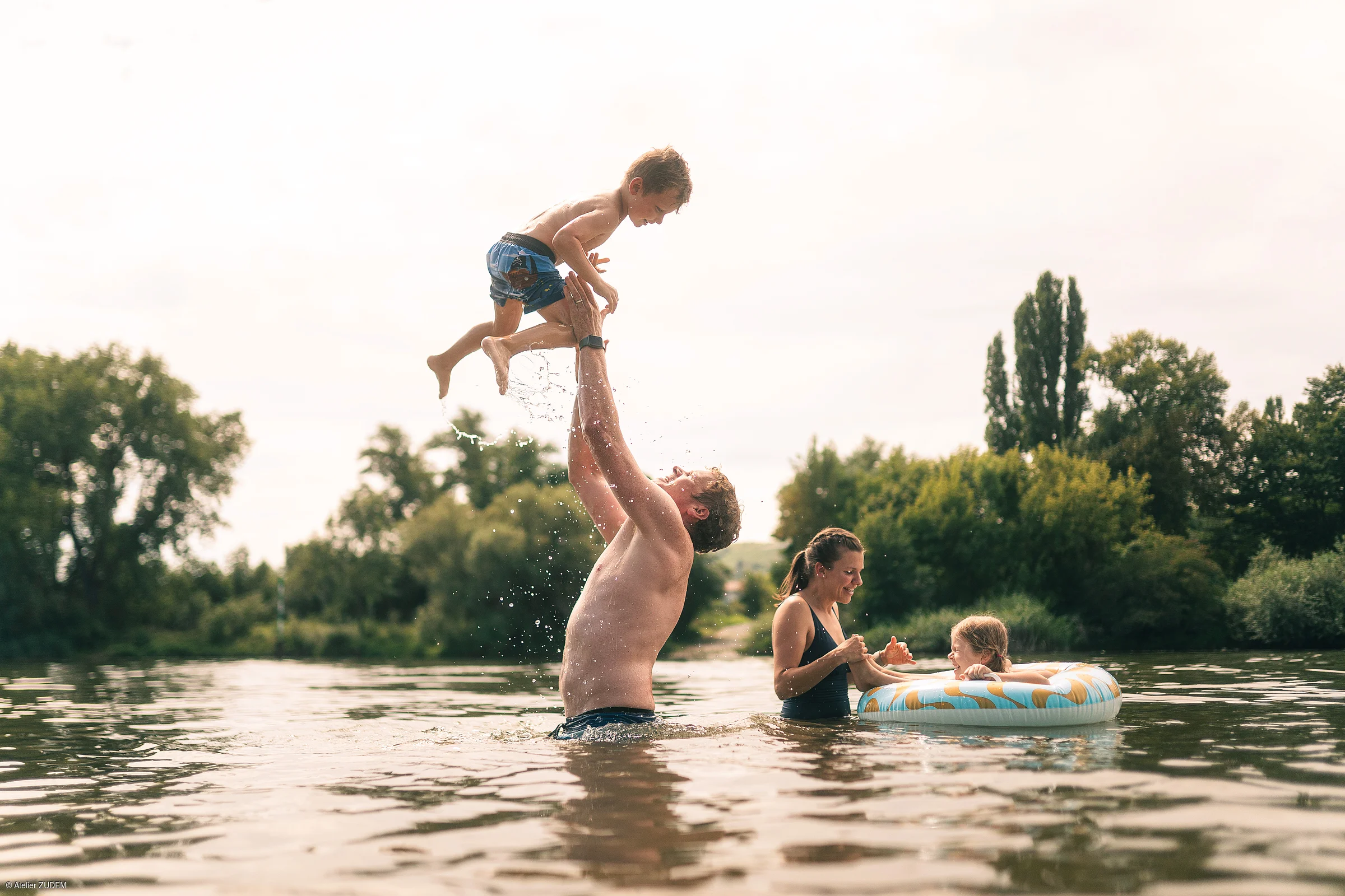 Vater wirft Kind in die Luft, Mutter hält anderes Kind im Schwimmreifen, alle im See, Bäume im Hintergrund.