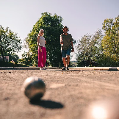 Zwei Personen spielen Boule auf einem Sandplatz im Freien, umgeben von Bäumen und Bänken.