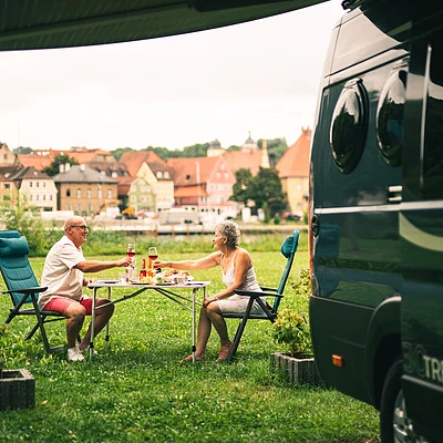 Älteres Paar sitzt an Campingtisch auf Wiese und stößt mit Weingläsern vor einem Wohnmobil an.