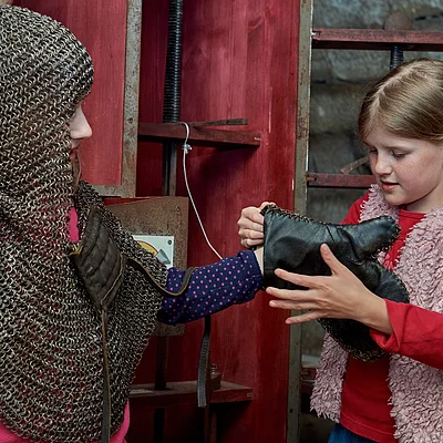 Zwei Mädchen, eines trägt Kettenhemd, das andere hilft beim Anziehen eines gepolsterten Handschuhs.