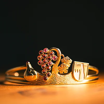 Goldener Armreif mit Trauben, Weinflasche, Weinblättern und Wappen auf Holztisch vor schwarzem Hintergrund