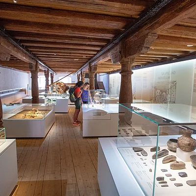 Museumsausstellung mit historischen Artefakten in Vitrinen und zwei Personen, die diese betrachten, Holzbalkendecke und Holzboden