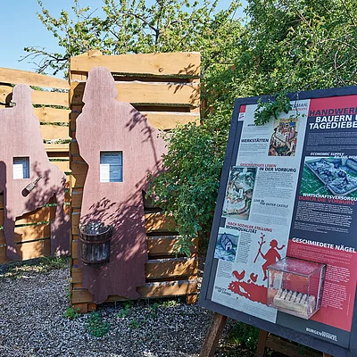 Informationsschild und zwei Holztafeln mit Figuren von Handwerkern im Freien bei Sonnenschein