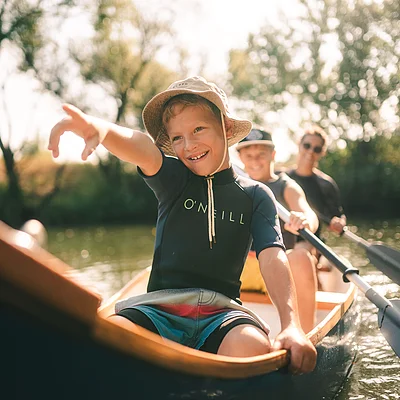 Drei Personen in einem Kanu auf einem Fluss, vorne ein Junge mit Sonnenhut, der nach vorne zeigt.