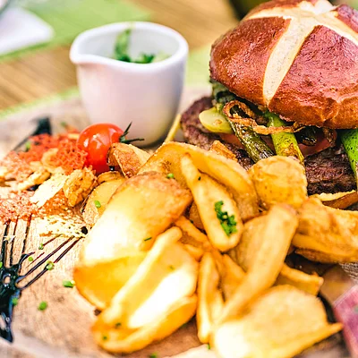 Holzbrett mit Burger, Kartoffelspalten, Kirschtomate und kleiner weißer Sauce-Schale, grüne Deko