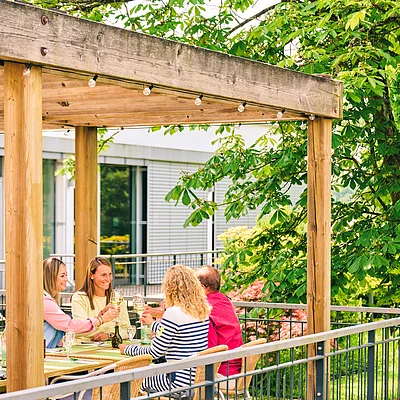 Vier Personen sitzen an einem Holztisch unter einem Holzpavillon und stoßen mit Weingläsern an.