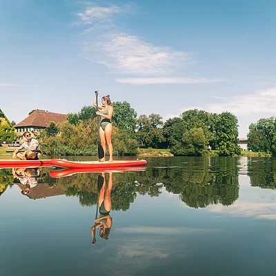 Zwei Personen auf roten Stand-Up-Paddle-Boards auf ruhigem Wasser vor Häusern und Bäumen bei klarem Himmel.