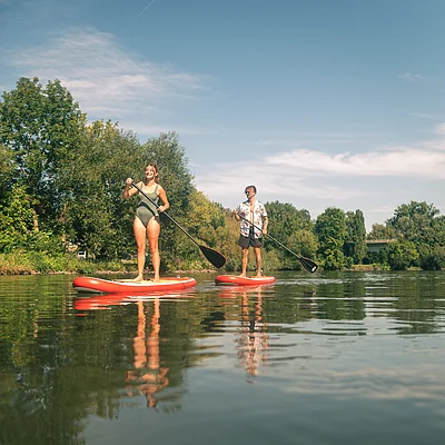 Zwei Personen stehen auf roten Stand-Up-Paddle-Boards auf einem ruhigen Fluss mit Bäumen am Ufer.
