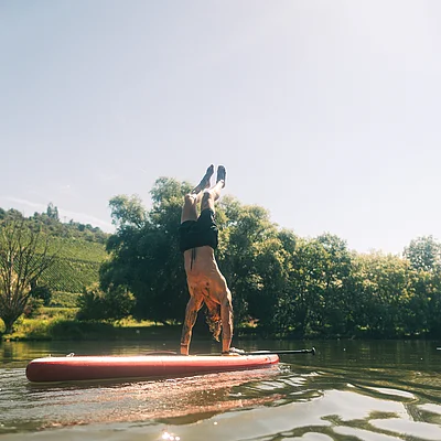 Mann macht Handstand auf einem roten Stand-up-Paddle-Board auf ruhigem Gewässer mit Bäumen im Hintergrund.