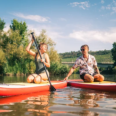 Zwei Personen sitzen auf Stand-up-Paddle-Boards auf einem ruhigen Fluss, umgeben von Bäumen und blauem Himmel.