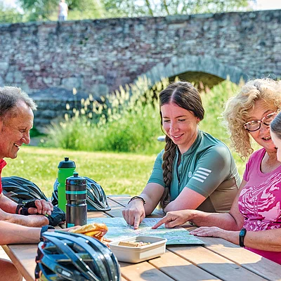 Fünf Personen sitzen an einem Holztisch im Freien und schauen gemeinsam auf eine Karte.