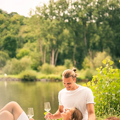 Paar liegt auf Picknickdecke am See, hält Weingläser, neben ihnen liegt ein brauner Hund.