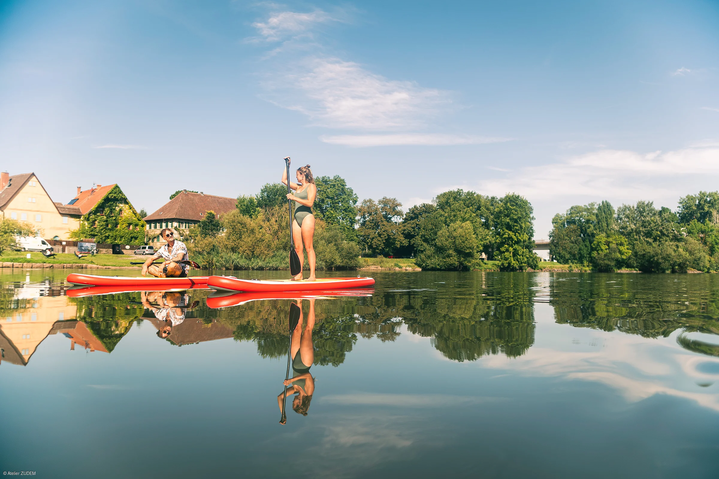Zwei Personen auf roten Stand-Up-Paddle-Boards auf ruhigem Wasser vor Häusern und Bäumen bei klarem Himmel.