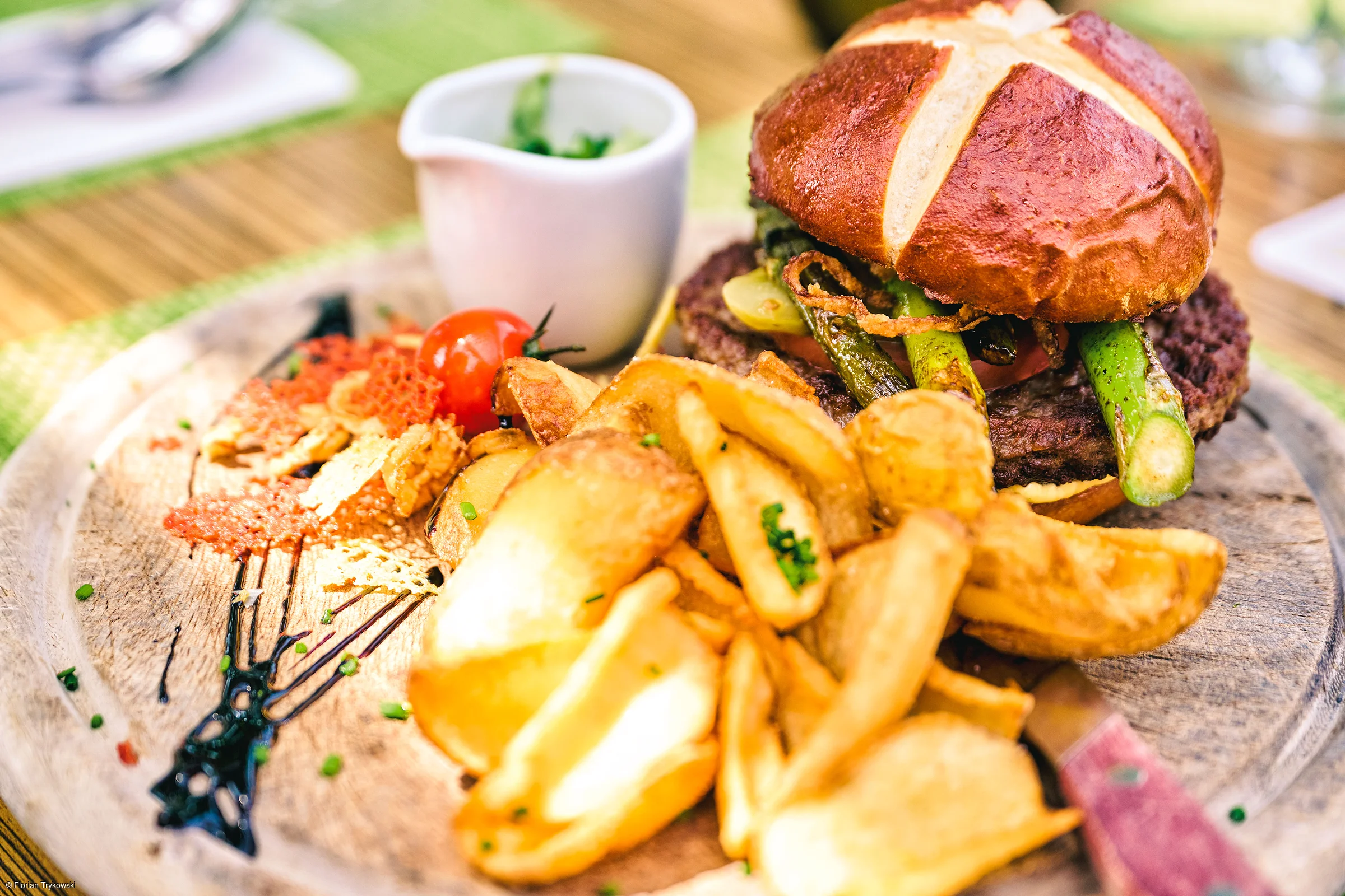 Holzbrett mit Burger, Kartoffelspalten, Kirschtomate und kleiner weißer Sauce-Schale, grüne Deko