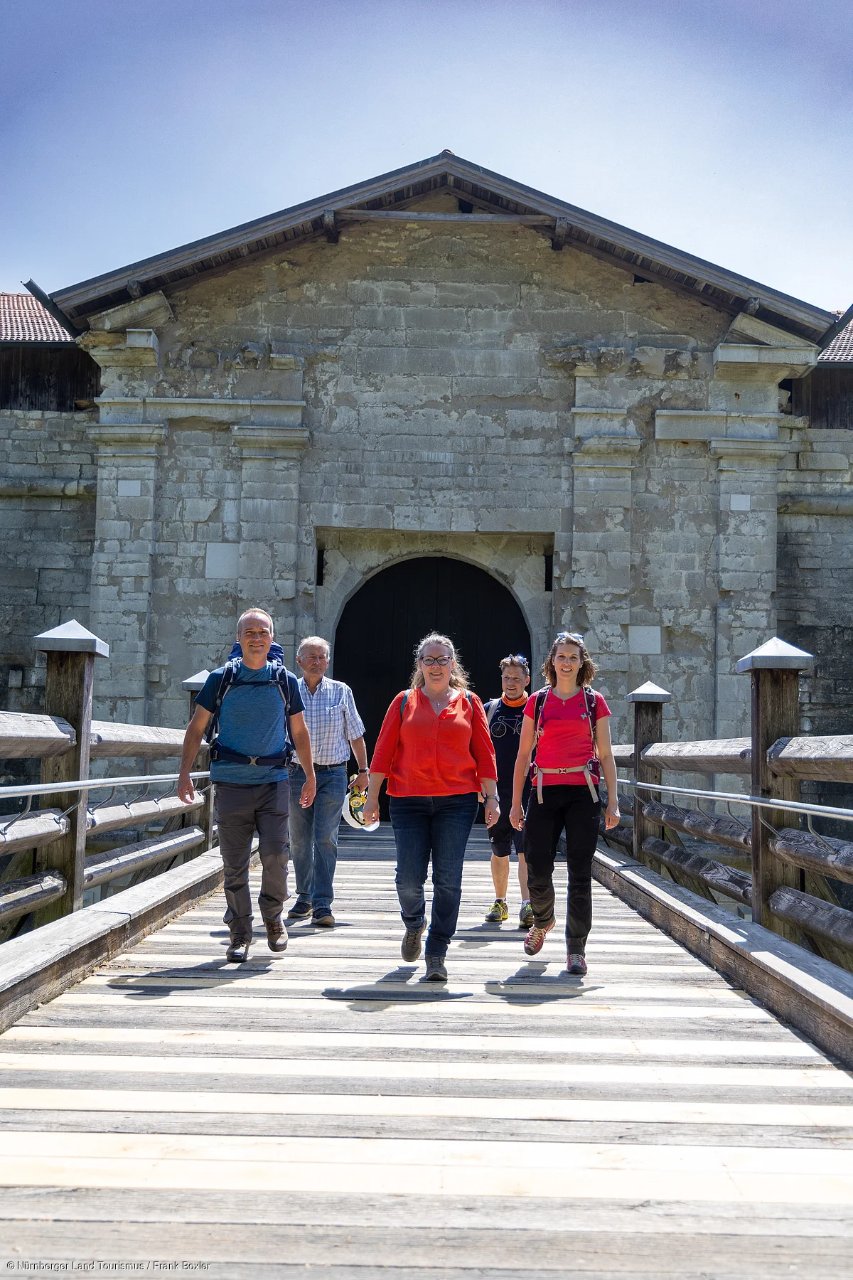 Fünf Personen mit Rucksäcken gehen auf einer Holzbrücke vor einem alten steinernen Torhaus bei Sonnenschein.