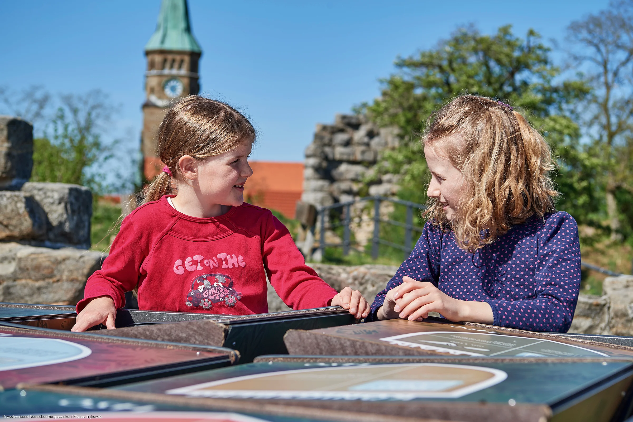 Zwei Mädchen sitzen draußen an einem Tisch mit Informationstafeln und schauen sich an.