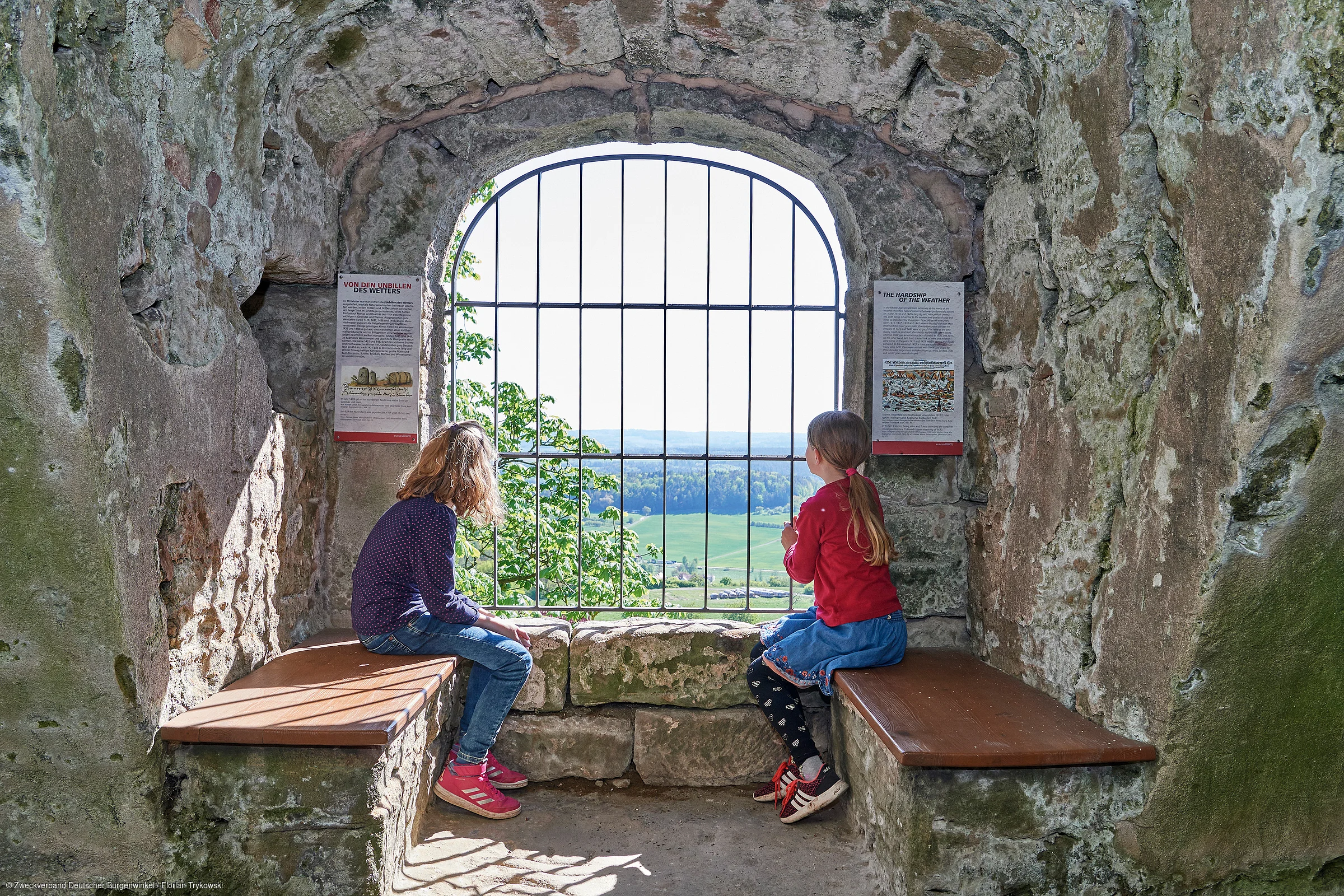 Zwei Kinder sitzen auf Steinbänken in einem steinernen Fenster mit Gitter und Blick auf Landschaft.
