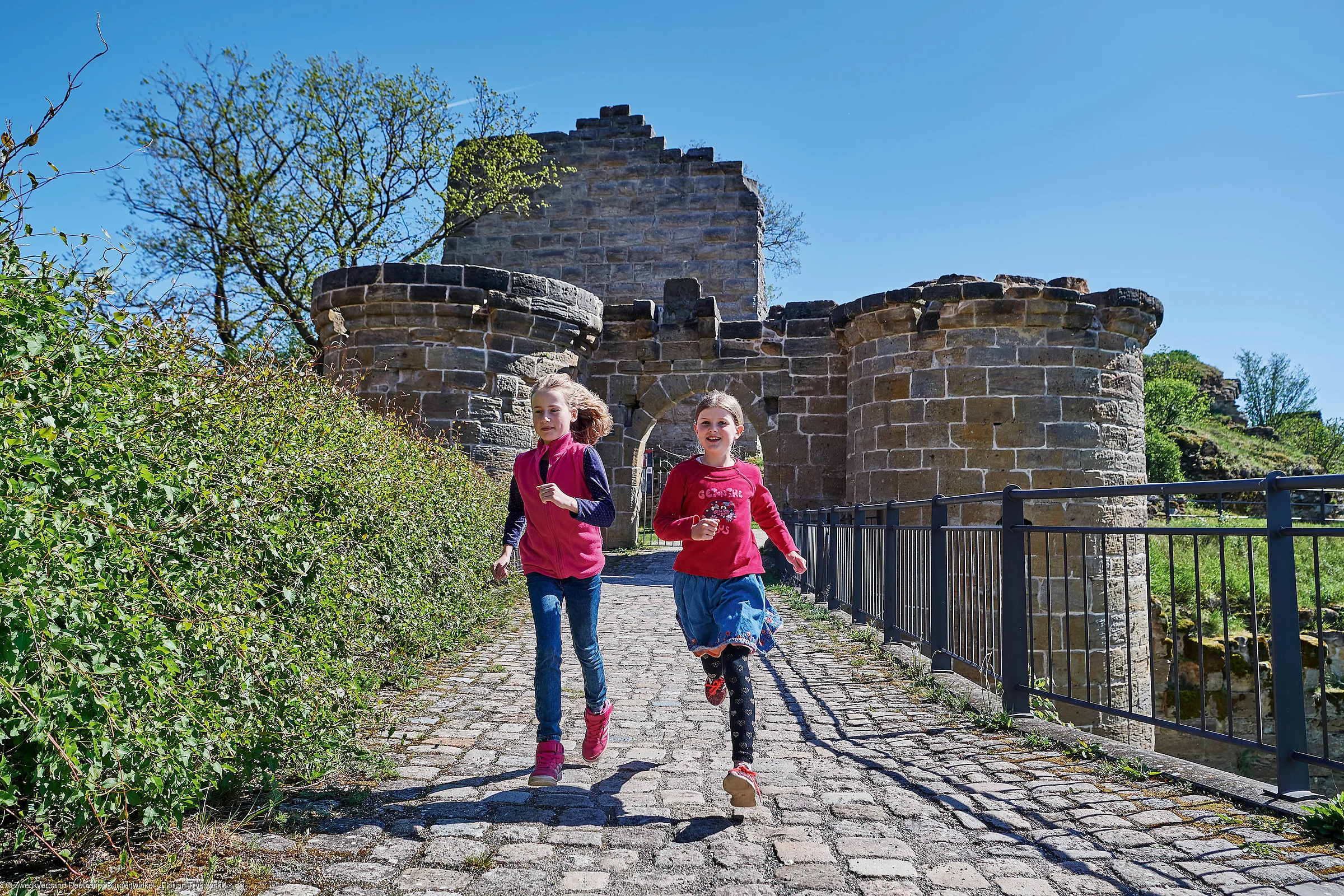 Zwei Mädchen laufen auf einem gepflasterten Weg vor einer alten Steinburg bei klarem Himmel.