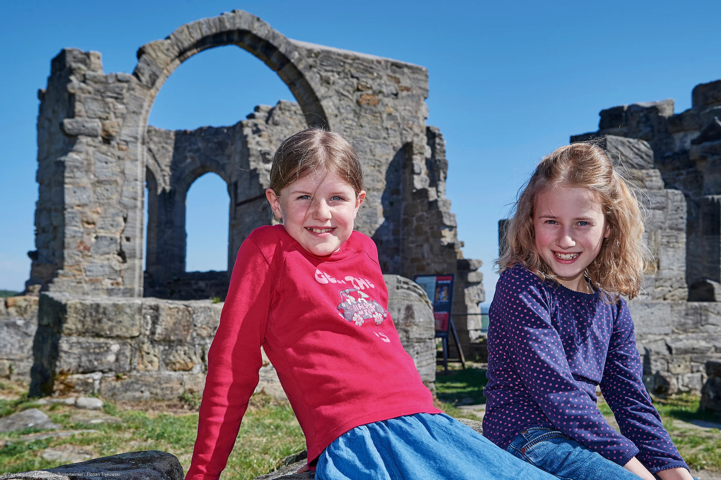 Zwei Mädchen sitzen vor einer alten Steinruine mit Bögen bei klarem Himmel.